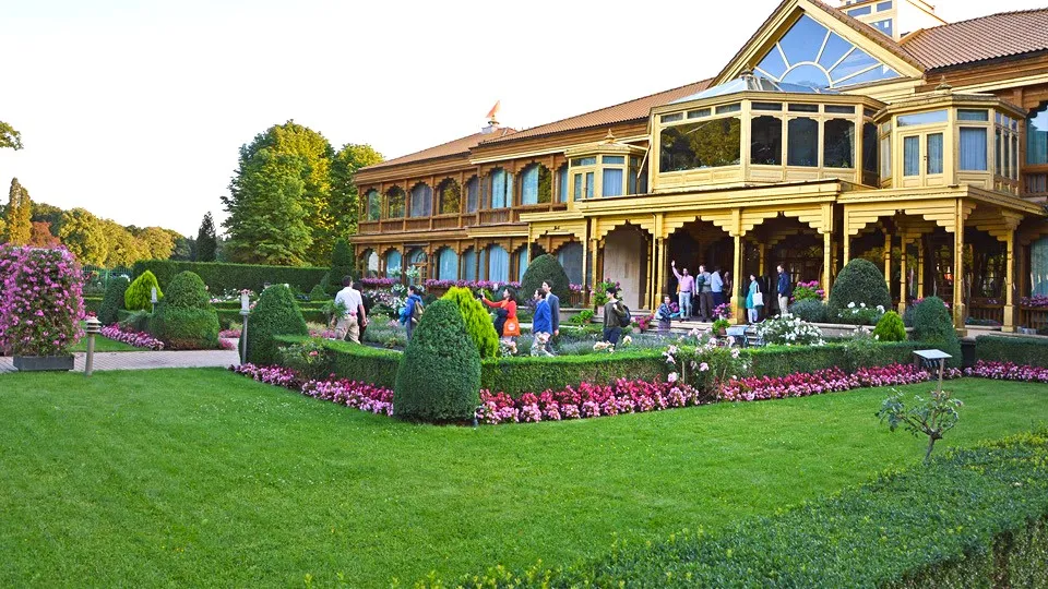 Aerial view of a grand ornate building with golden-brown facade surrounded by meticulously maintained gardens with manicured lawns, shaped hedges, and vibrant flower beds under a clear sky