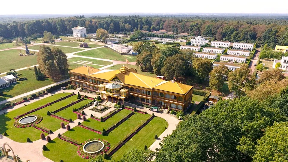 Aerial view of a large opulent estate featuring a grand central building with golden-yellow multi-tiered roof, elaborate formal gardens with fountains, and an array of white classical and modern buildings nestled within a vast forest